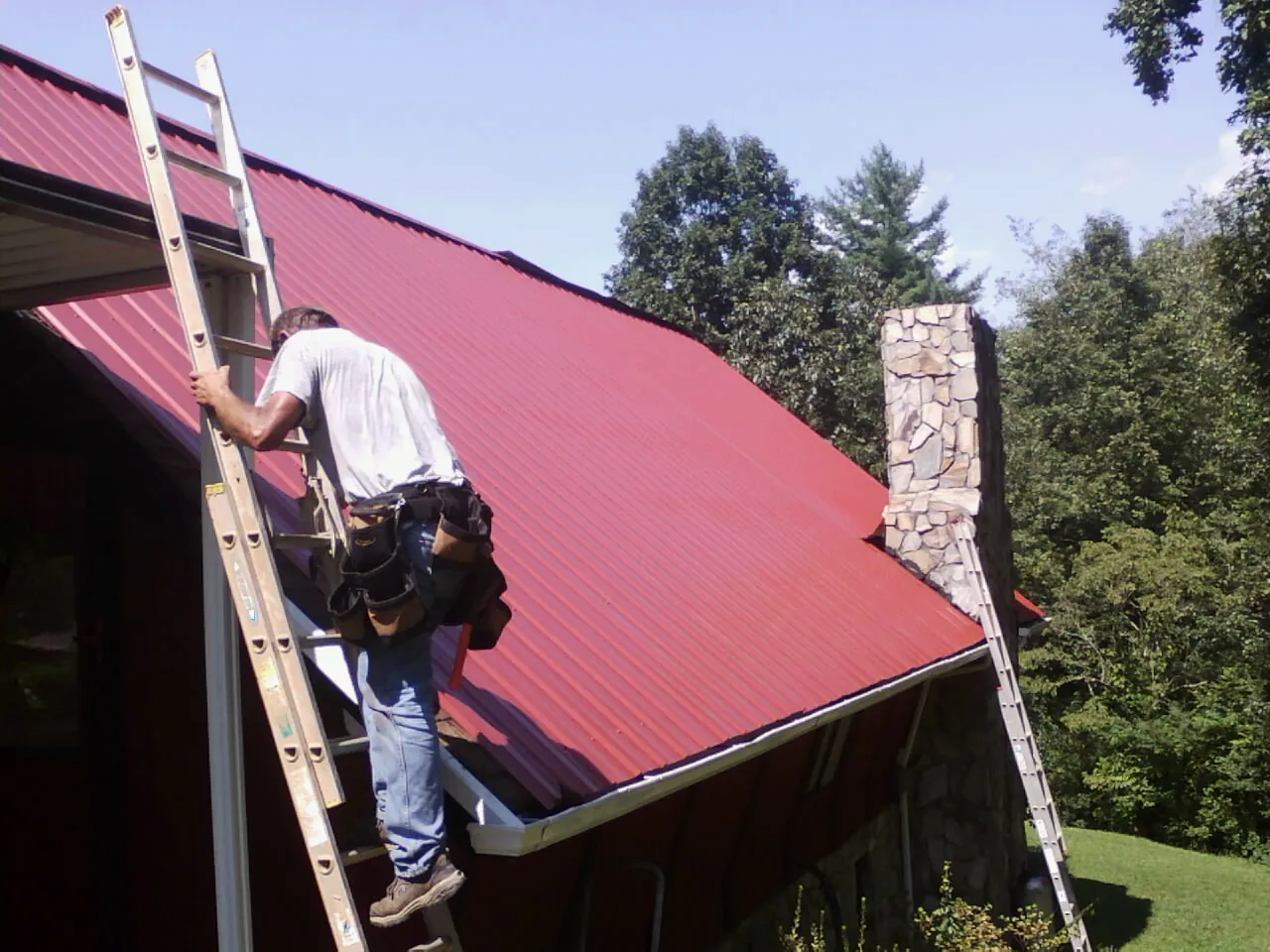 Roofer on a ladder beside a red metal roof and stone chimney — Soffit & Fascia Repair by Roofing Brothers in San Pedro
